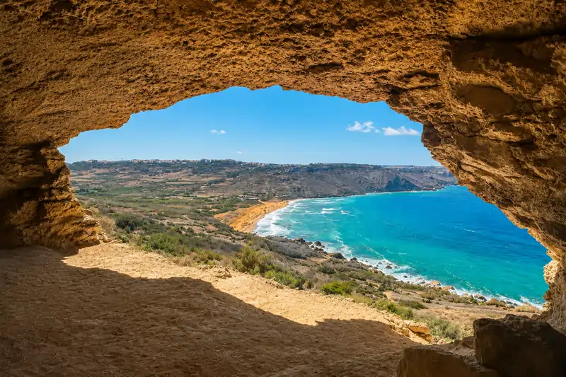 Una bahía turquesa y laderas en terrazas vistas desde la boca de una cueva de piedra caliza en la costa de Gozo.
